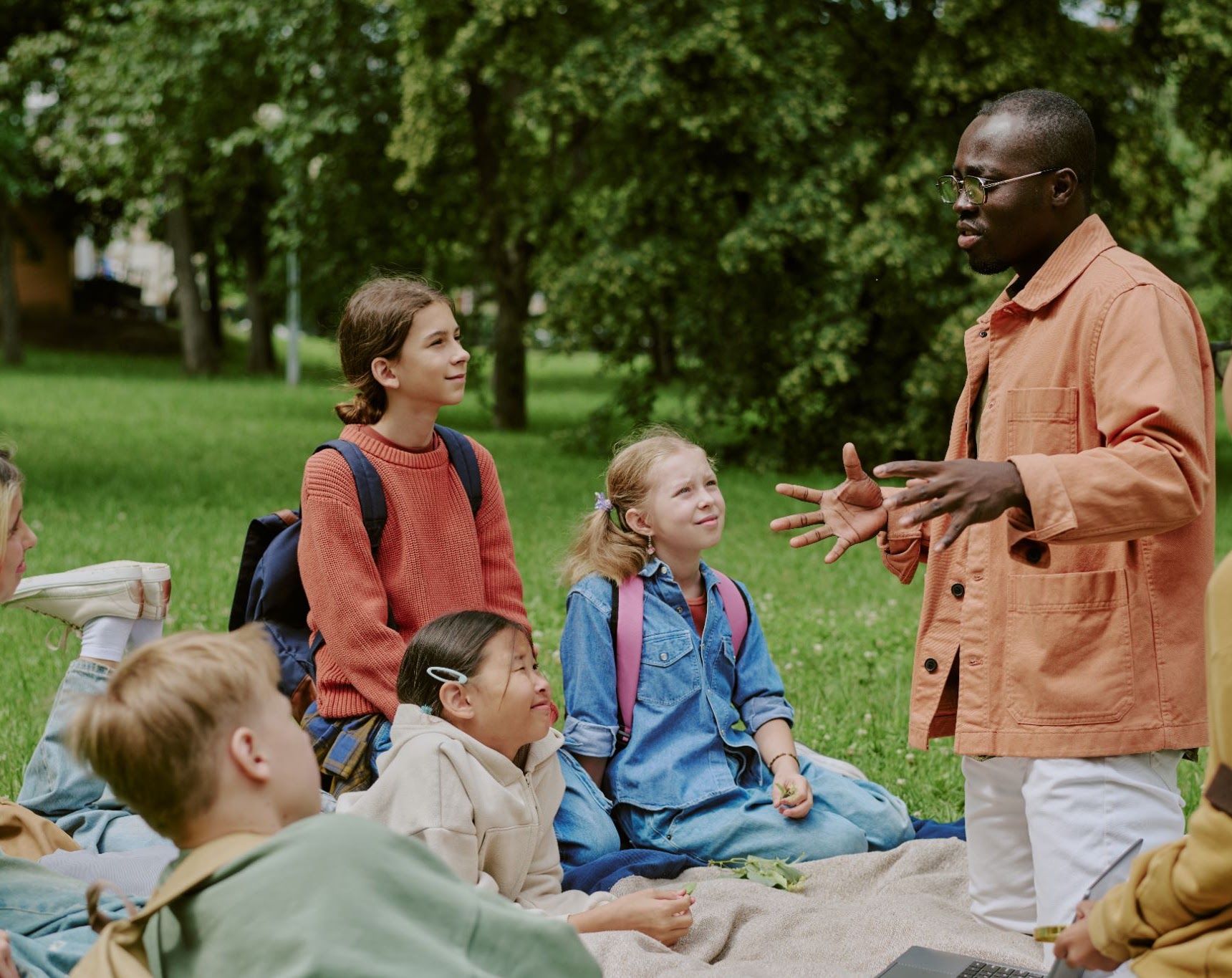 An adult talking with a group of children seated on the grass in a park.